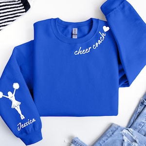 May include: Royal blue sweatshirt with white text and graphics. The text "cheer coach" is on the chest, and a cheerleader graphic with the name "Jessica" is on the sleeve. A white heart is next to the text "cheer coach".