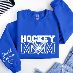 Pu&ograve; includere: Felpa blu reale con testo bianco che recita "Hockey Mom" con un design di bastone da hockey e disco. La manica sinistra ha un testo bianco che recita "David Jackson" con un cuore.