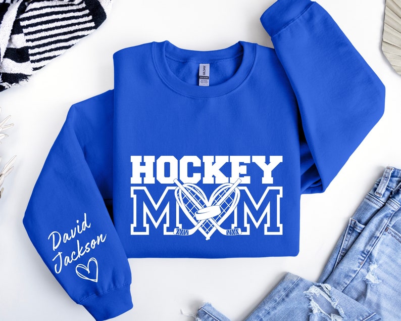 Pu&ograve; includere: Felpa blu reale con testo bianco che recita "Hockey Mom" con un design di bastone da hockey e disco. La manica sinistra ha un testo bianco che recita "David Jackson" con un cuore.