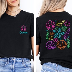 May include: Black t-shirt with a seashell design and the name "Jessica" in teal. The back of the shirt features a colorful neon design of seashells, starfish, and flowers. The shirt is made of a soft material.