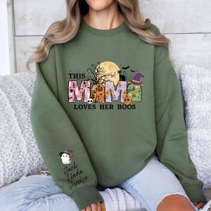May include: A green sweatshirt with a Halloween-themed graphic that says "This Mama Loves Her Boos". The graphic features a moon, bats, a witch's hat, and a black cat. The sweatshirt has a crew neck and long sleeves.