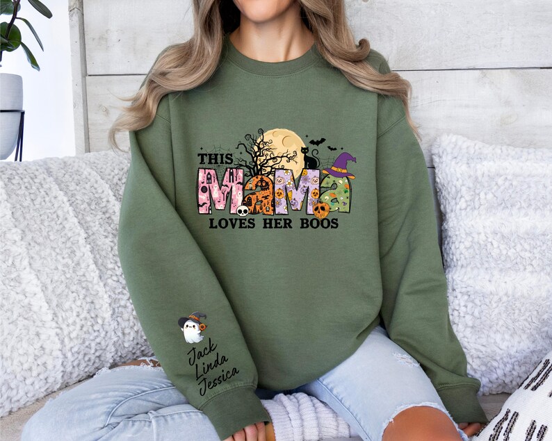 May include: A green sweatshirt with a Halloween-themed graphic that says "This Mama Loves Her Boos". The graphic features a moon, bats, a witch's hat, and a black cat. The sweatshirt has a crew neck and long sleeves.