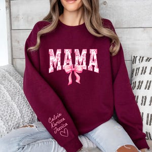 May include: Burgundy sweatshirt with the word "MAMA" in white letters with pink floral accents. The sweatshirt has a pink bow and the names "Calvin, Karissa, Jessica" written in white on the sleeve.