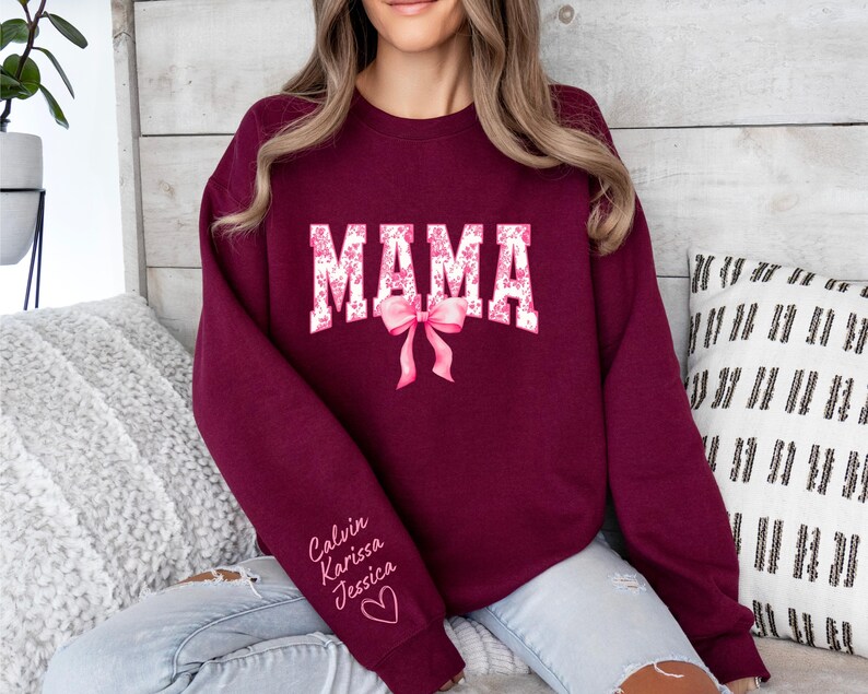 May include: Burgundy sweatshirt with the word "MAMA" in white letters with pink floral accents. The sweatshirt has a pink bow and the names "Calvin, Karissa, Jessica" written in white on the sleeve.