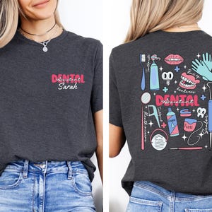 May include: A dark grey t-shirt with a pink and white design that says "Dental Assistant Sarah" on the front and a colourful illustration of dental tools and equipment on the back.