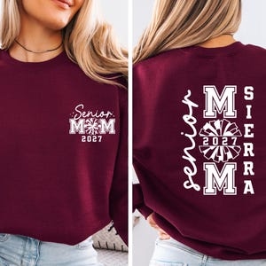 May include: A maroon sweatshirt with white text. The front features "Senior Mom 2027" with a cheerleader graphic. The back displays "Senior Mom" vertically, with "Sierra" and "2027" and a cheerleader graphic.