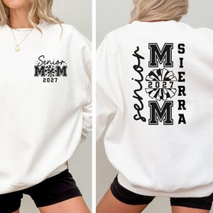 May include: White sweatshirt with black text and graphics. The front features "Senior Mom 2027" with a pom-pom design. The back displays "Senior" vertically, and "Sierra Mom 2027" with a pom-pom graphic.