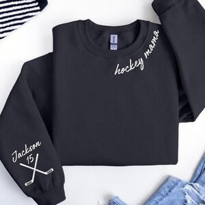 May include: Black crewneck sweatshirt with the words "hockey mama" in white script. The left sleeve has the name "Jackson" and the number "15" with crossed hockey sticks. The sweatshirt is a casual apparel item.