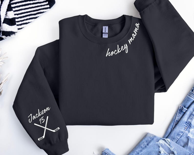 May include: Black crewneck sweatshirt with the words "hockey mama" in white script. The left sleeve has the name "Jackson" and the number "15" with crossed hockey sticks. The sweatshirt is a casual apparel item.