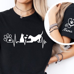 May include: Black t-shirt with a white graphic of a cat and dog with a heartbeat line and paw print. The shirt also has a stethoscope with a paw print on the sleeve and the word "Name" underneath.