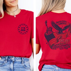 Pode incluir: Camiseta vermelha com um gr&aacute;fico circular azul marinho na frente que diz "1776 2026". A parte de tr&aacute;s apresenta um design com a Est&aacute;tua da Liberdade, uma &aacute;guia e uma faixa que diz "United States of America 250th Anniversary 1776-2026".