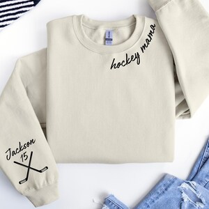 May include: Cream-colored sweatshirt with the words "hockey mama" in black script on the upper chest. The left sleeve features the name "Jackson", the number "15", and crossed hockey sticks. The sweatshirt is folded on a white surface.