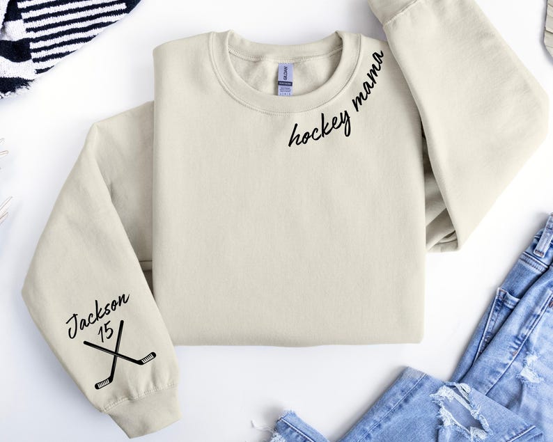 May include: Cream-colored sweatshirt with the words "hockey mama" in black script on the upper chest. The left sleeve features the name "Jackson", the number "15", and crossed hockey sticks. The sweatshirt is folded on a white surface.