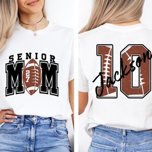 May include: A white t-shirt featuring a front graphic with the text SENIOR MOM and a football with the number 27. The back graphic displays the number 10 with a football texture and the name Jackson written in black script across the number.