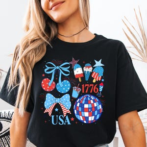 Puede incluir: Camiseta negra con un dise&ntilde;o patri&oacute;tico en rojo, blanco y azul. El gr&aacute;fico incluye lazos, helados, una bola de discoteca y los textos "USA" y "1776".