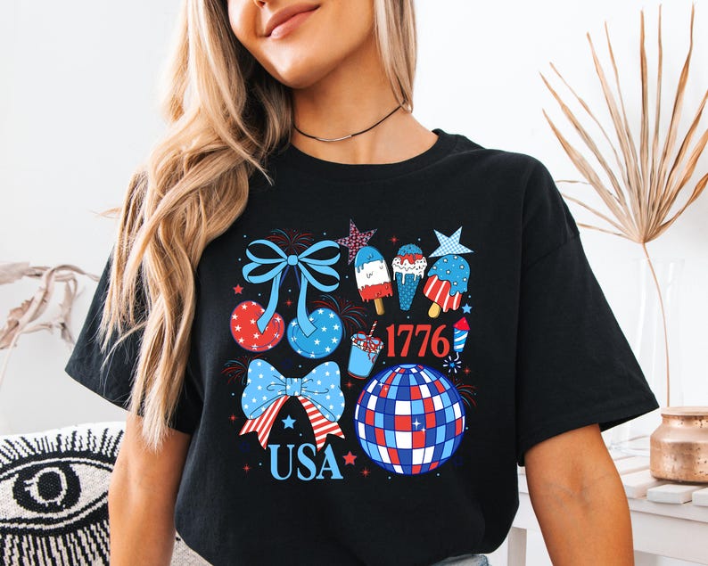 Puede incluir: Camiseta negra con un dise&ntilde;o patri&oacute;tico en rojo, blanco y azul. El gr&aacute;fico incluye lazos, helados, una bola de discoteca y los textos "USA" y "1776".