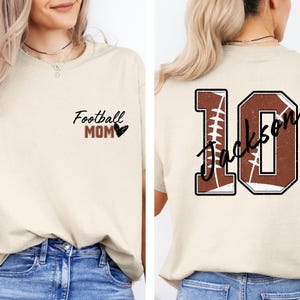 May include: A beige t-shirt with the text "Football Mom" and a heart on the front. The back of the shirt has a brown and white football design with the number 10 and the text "Jackson" on it.