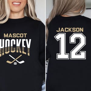 K&ouml;nnte beinhalten: Schwarzer Sweatshirt mit goldener Aufschrift "Mascot Hockey" und einer Grafik von zwei gekreuzten Hockeyschl&auml;gern &uuml;ber einem Puck. Die R&uuml;ckseite des Sweatshirts hat wei&szlig;en Text mit der Aufschrift "Jackson 12".