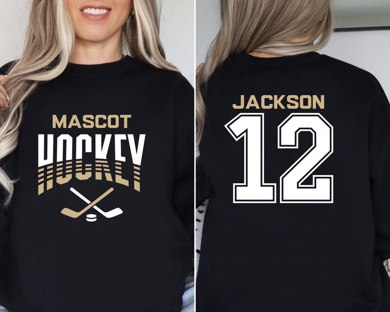 K&ouml;nnte beinhalten: Schwarzer Sweatshirt mit goldener Aufschrift "Mascot Hockey" und einer Grafik von zwei gekreuzten Hockeyschl&auml;gern &uuml;ber einem Puck. Die R&uuml;ckseite des Sweatshirts hat wei&szlig;en Text mit der Aufschrift "Jackson 12".