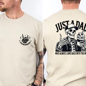 May include: Beige t-shirt with a skeleton graphic. The front features a skeleton hand design with the word "Fatherhood." The back displays a skeleton wearing a cap and holding a milk carton, with the text "Just a Dad Who Always Came Back With The Milk."