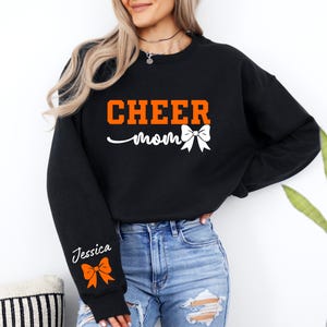 May include: Black sweatshirt with the text "CHEER mom" in orange and white. The sweatshirt has a white bow on the front and a personalized name on the sleeve with an orange bow.
