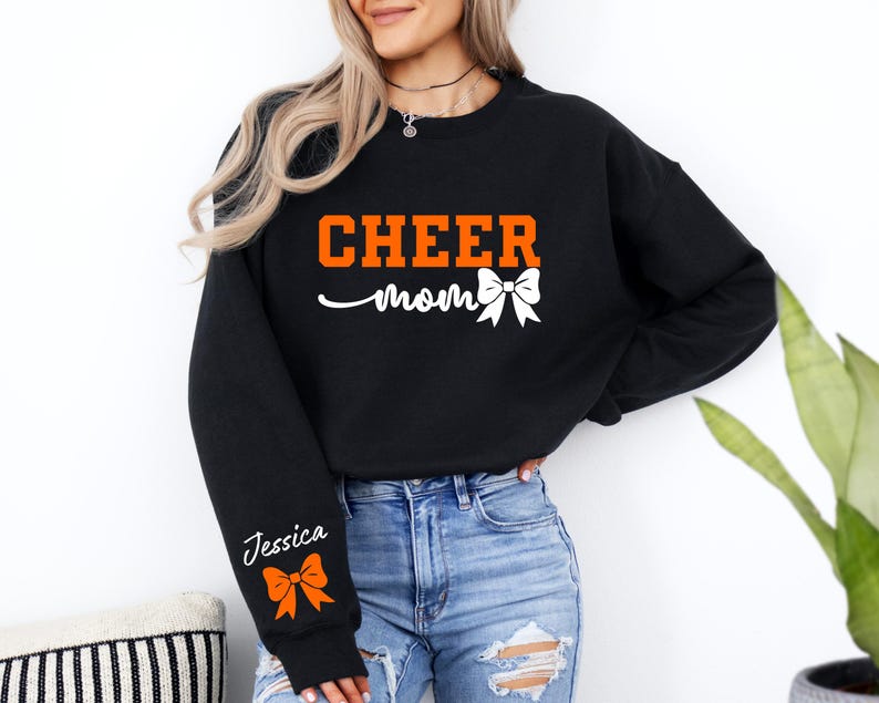 May include: Black sweatshirt with the text "CHEER mom" in orange and white. The sweatshirt has a white bow on the front and a personalized name on the sleeve with an orange bow.
