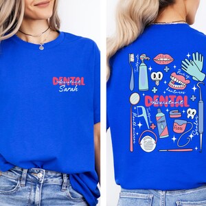 May include: A royal blue t-shirt with a dental assistant design on the back. The design features a variety of dental tools, including a toothbrush, toothpaste, floss, and a mouth mirror. The text "Dental Assistant" is written in pink and white letters. The front of the shirt has the text "Dental Assistant Sarah" in pink and white letters.