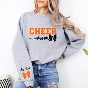 May include: Gray sweatshirt with the text "CHEER mom" in orange and a black bow. The left sleeve has the text "Jessica" in black with an orange bow.