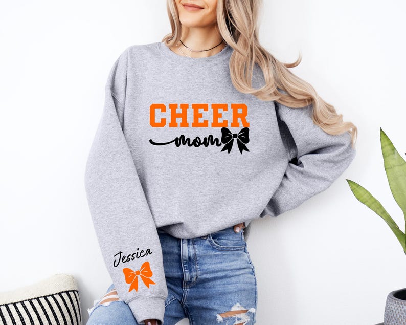 May include: Gray sweatshirt with the text "CHEER mom" in orange and a black bow. The left sleeve has the text "Jessica" in black with an orange bow.