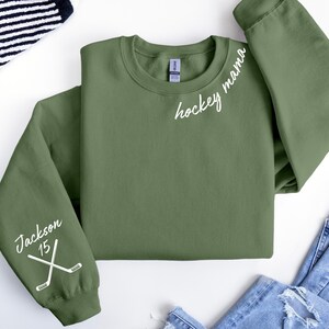 May include: Olive green sweatshirt with the words "hockey mama" in white script. The left sleeve features the name "Jackson" and the number "15" with crossed hockey sticks. The sweatshirt is laid flat on a white surface.
