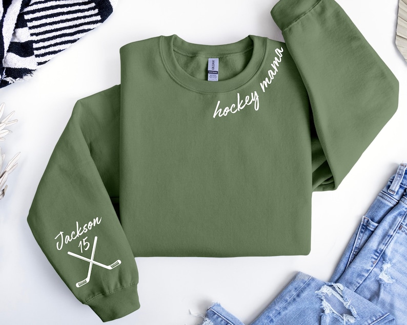 May include: Olive green sweatshirt with the words "hockey mama" in white script. The left sleeve features the name "Jackson" and the number "15" with crossed hockey sticks. The sweatshirt is laid flat on a white surface.