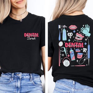 May include: Black t-shirt with a pink and blue graphic of dental tools and the text "Dental Assistant Sarah" on the front. The back of the shirt has a pink and blue graphic of dental tools and the text "Dental Assistant".