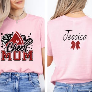Puede incluir: Camiseta rosa de mam&aacute; de animadora con un lazo de animadora rojo con purpurina, pompones y el texto "Cheer Mom". La parte posterior de la camiseta tiene un lazo rojo con purpurina y el nombre "Jessica" en negro.