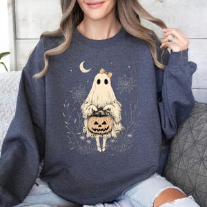 May include: A navy blue sweatshirt with a white ghost holding a jack-o'-lantern. The ghost is wearing a pink bow and is surrounded by spiderwebs and leaves.