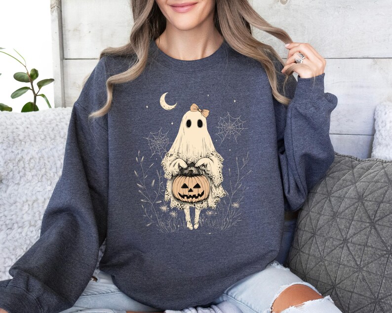 May include: A navy blue sweatshirt with a white ghost holding a jack-o'-lantern. The ghost is wearing a pink bow and is surrounded by spiderwebs and leaves.