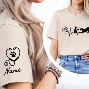 May include: Two beige t-shirts with a black paw print design. One shirt has a stethoscope with a paw print in the heart. The other shirt has a paw print, a cat, and a dog with a heartbeat line. The text "Name" is on the shirt with the stethoscope.
