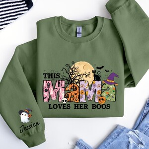 May include: Green sweatshirt with a Halloween-themed design that says "This Mama Loves Her Boos". The design features a black cat, a witch's hat, a moon, and a ghost with the name "Jessica" on the sleeve.