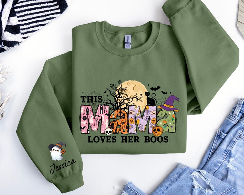 May include: Green sweatshirt with a Halloween-themed design that says "This Mama Loves Her Boos". The design features a black cat, a witch's hat, a moon, and a ghost with the name "Jessica" on the sleeve.
