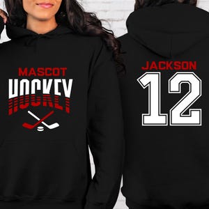 K&ouml;nnte beinhalten: Schwarzer Hoodie mit einem wei&szlig;en und roten Hockey-Design auf der Vorderseite. Der Text auf der Vorderseite lautet "Mascot Hockey". Die R&uuml;ckseite des Hoodies hat eine wei&szlig;e Nummer "12" mit roten Umrandungen und den Text "Jackson" &uuml;ber der Nummer.