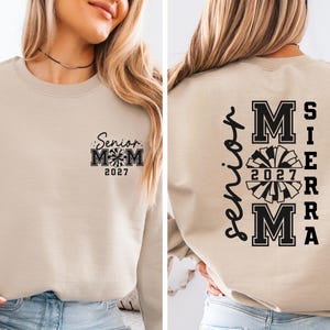 May include: Beige sweatshirt with black text and graphics. The front features "Senior Mom 2027" with a pom-pom graphic. The back displays "Senior" vertically, and "MOM SIERRA 2027" with a pom-pom graphic.