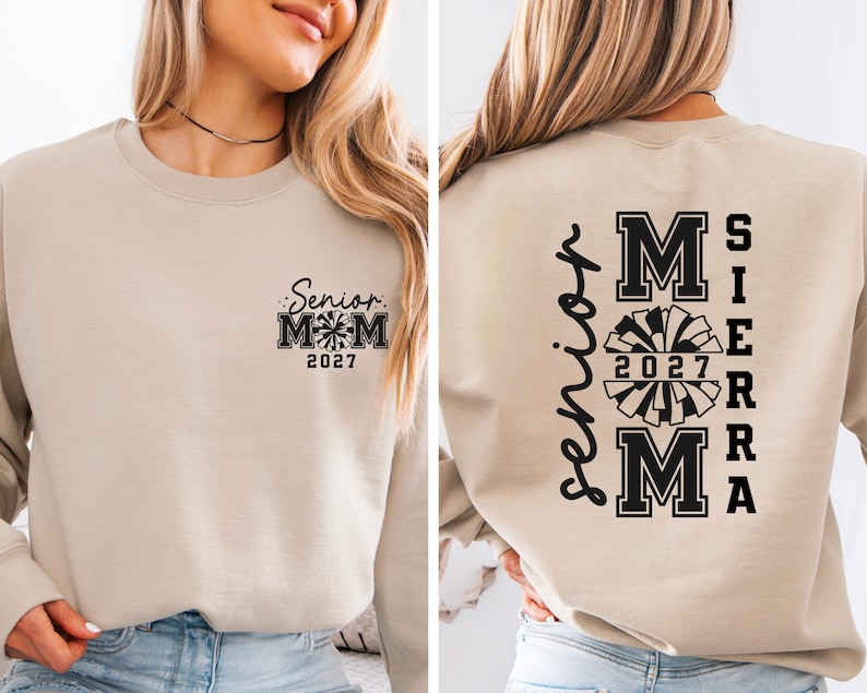 May include: Beige sweatshirt with black text and graphics. The front features "Senior Mom 2027" with a pom-pom graphic. The back displays "Senior" vertically, and "MOM SIERRA 2027" with a pom-pom graphic.