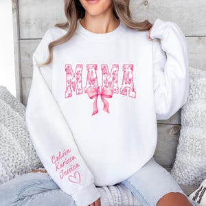May include: White sweatshirt with the word "MAMA" in pink with a floral pattern. The word is in a large, bold font. A pink bow is below the word. The sleeve has the names "Calvin, Karissa, Jessica" written in pink.