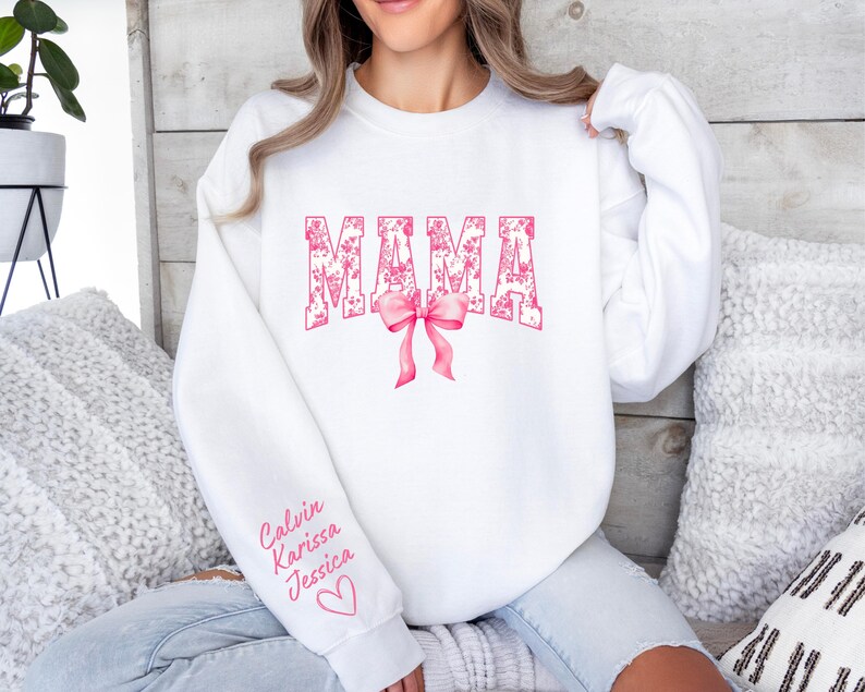 May include: White sweatshirt with the word "MAMA" in pink with a floral pattern. The word is in a large, bold font. A pink bow is below the word. The sleeve has the names "Calvin, Karissa, Jessica" written in pink.