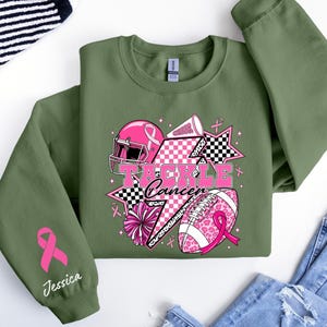 Op de afbeelding: Olijfgroene sweatshirt met een roze en zwarte afbeelding met de tekst "Tackle Cancer". De afbeelding bevat een footballhelm, football, sterren en cheerleading-elementen. Een roze lint en de naam "Jessica" staan op de linkermouw.