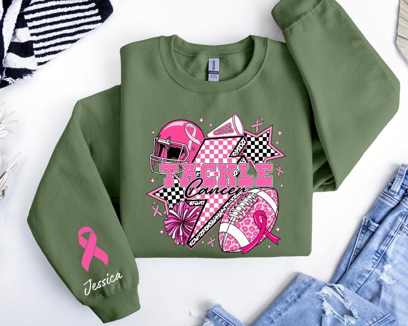 Op de afbeelding: Olijfgroene sweatshirt met een roze en zwarte afbeelding met de tekst "Tackle Cancer". De afbeelding bevat een footballhelm, football, sterren en cheerleading-elementen. Een roze lint en de naam "Jessica" staan op de linkermouw.