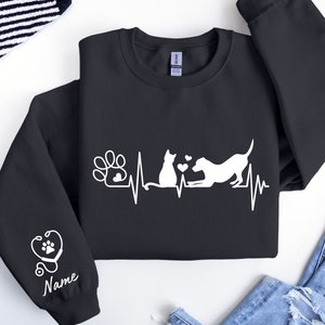 May include: Black sweatshirt with a white design of a cat and dog with a heartbeat line and paw print. The sleeve has a white design of a stethoscope and heart with the word "Name" underneath.