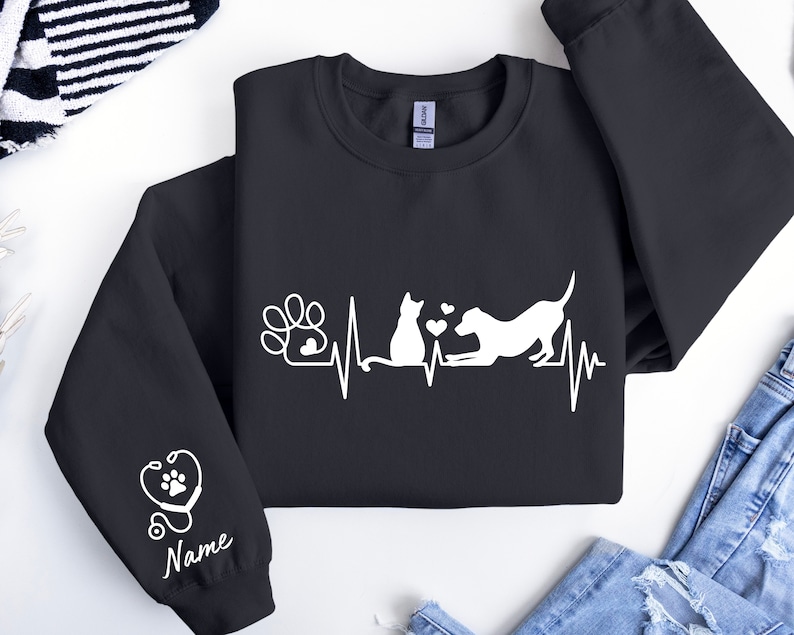 May include: Black sweatshirt with a white design of a cat and dog with a heartbeat line and paw print. The sleeve has a white design of a stethoscope and heart with the word "Name" underneath.