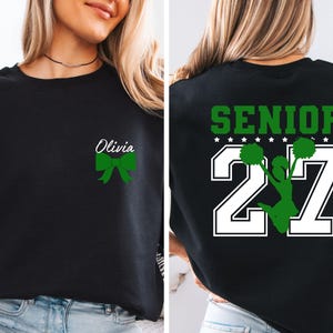 May include: A black crewneck sweatshirt featuring a small green bow and the name Olivia on the front left chest. The back displays the word SENIOR in green above the number 27 in white, with a green silhouette of a cheerleader holding pom-poms.