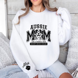 May include: White sweatshirt with black text that reads "Aussie Mom Like a Normal Mom Except Much Cooler" and a black and white image of an Australian Shepherd dog. The left sleeve has black text that reads "Alex" with a paw print.