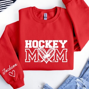 Pu&ograve; includere: Felpa rossa con testo bianco che dice "Hockey Mom" con un bastone da hockey e un disco a forma di cuore. La manica sinistra ha il nome "Jackson" scritto in corsivo con un cuore.